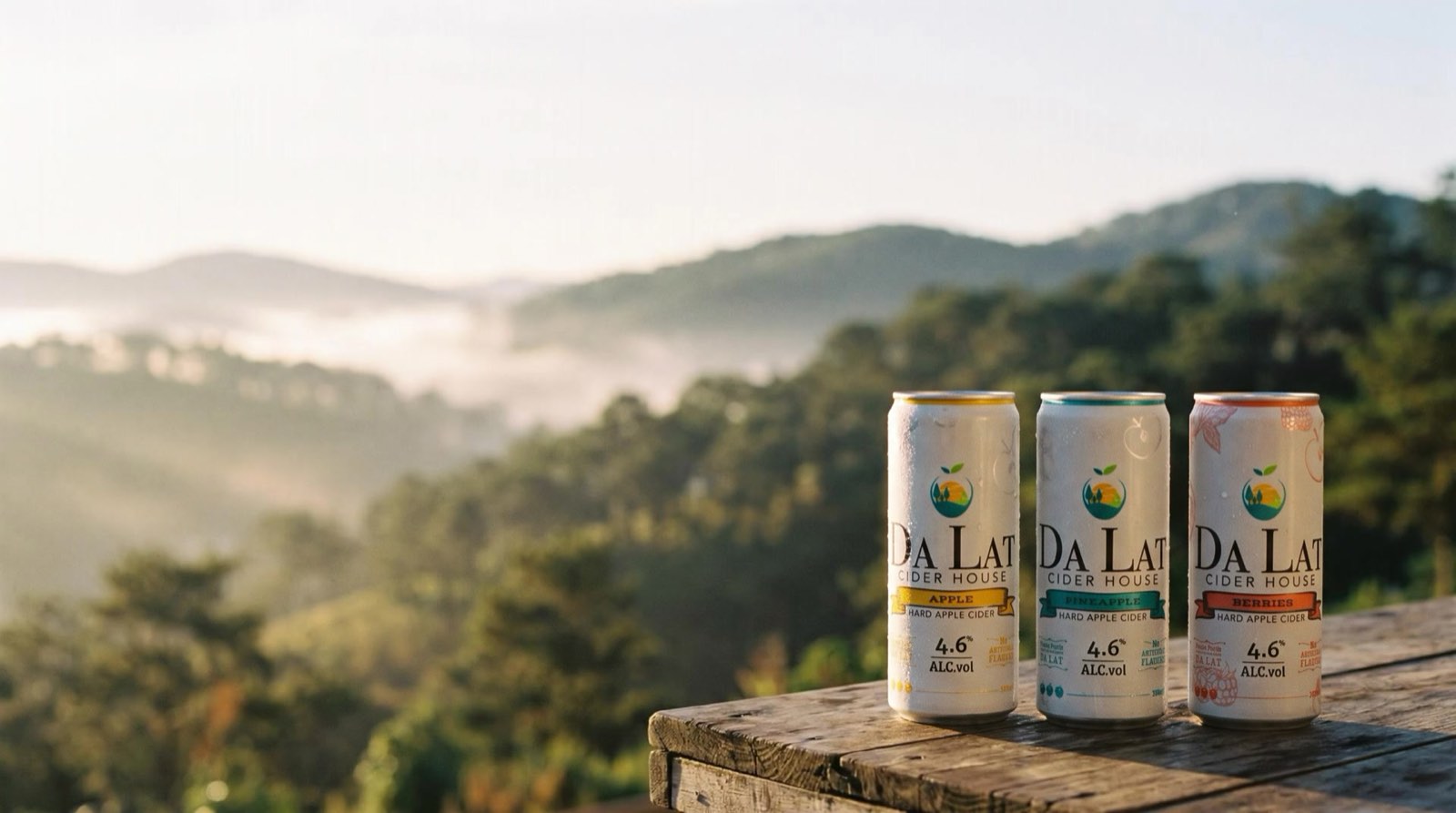 Three Da Lat Cider cans on a wooden veranda table above misty pine hills